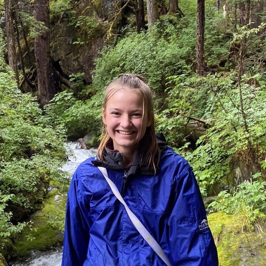 Angling Unlimited crew member, Addison Lindow, smiles in the forest wearing a blue raincoat