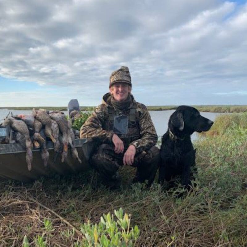 Angling Unlimited crew member, Hayden Bishop poses next to a black dog and a boat with hunted ducks